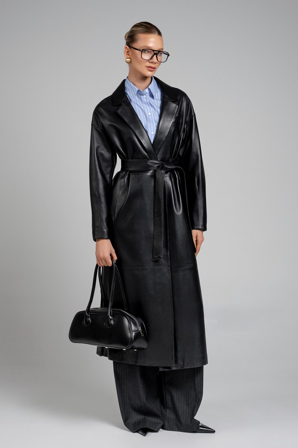 A woman wears a bold black leather coat with wide, structured shoulders, paired with wide trousers and a sleek black fashion leather bag, exuding modern power dressing.