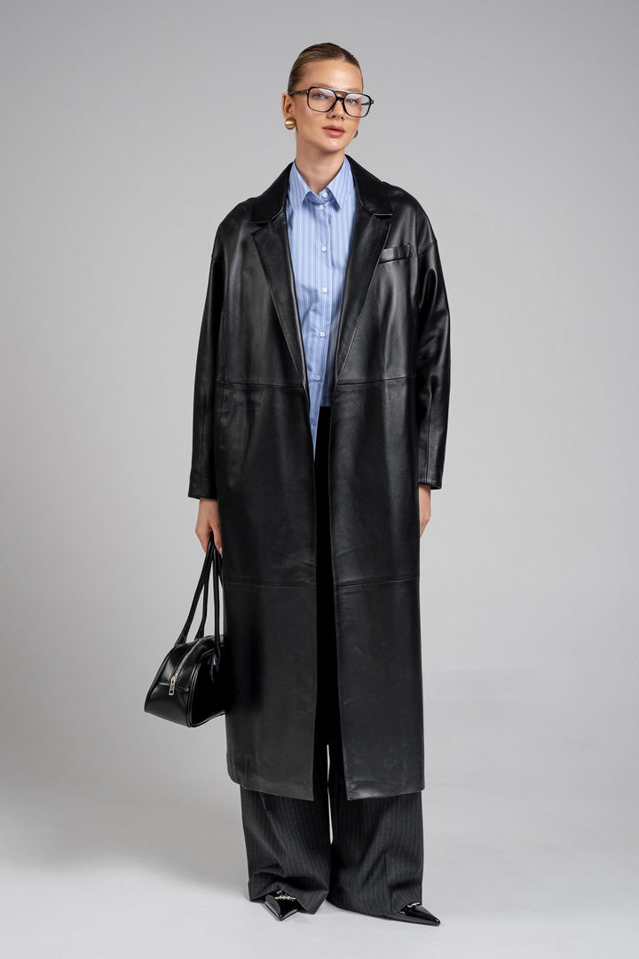 Wearing wide-legged trousers and a fashion-forward black leather handbag, a woman stuns in a structured genuine leather coat with strong shoulders and refined tailoring.