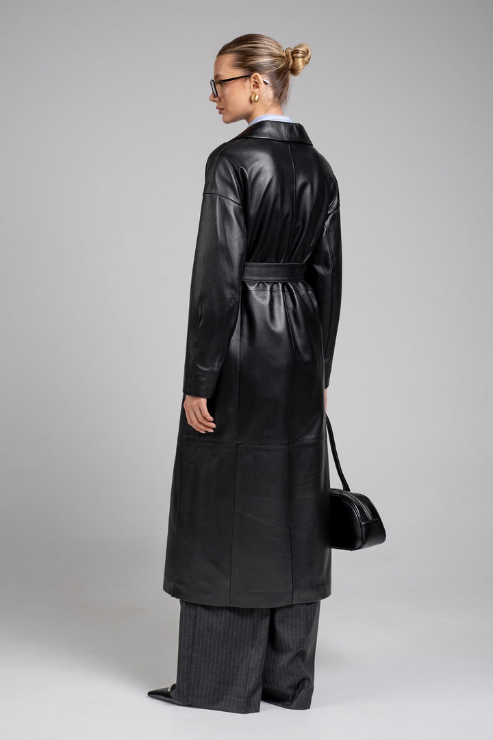 A woman wears a sharply tailored black leather coat featuring wide shoulders and a cinched waist, styled with elegant wide trousers and a chic black leather bag.