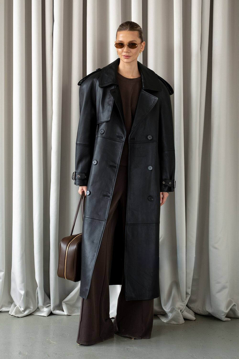 Elegantly dressed, a woman models a black leather trench coat with a classic double-breasted design, sophisticated notched lapels, and belted cuffs made from premium soft leather.