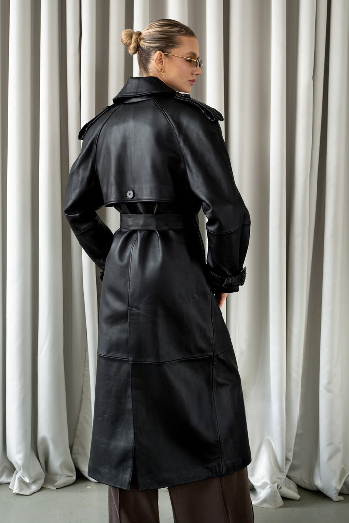 A woman is seen wearing a genuine black leather trench coat, combining timeless double-breasted style with buttery soft leather and refined belted cuff details.